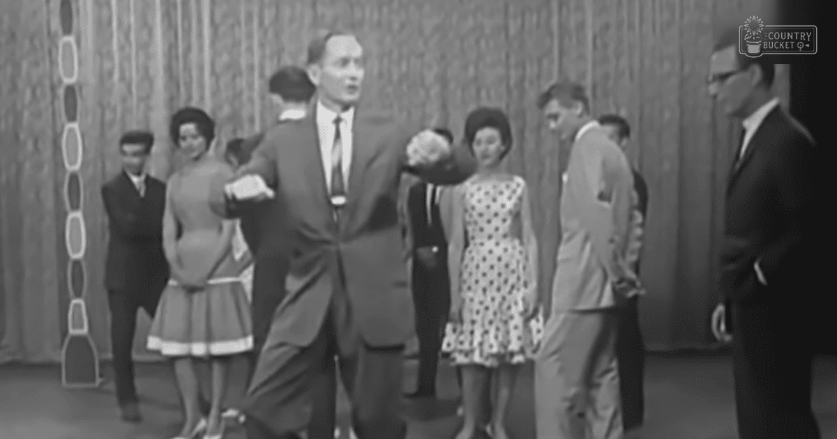 Adult male in motion possibly dancing indoors at an event or social gathering with others standing around him; a curtain with vertical stripes and circular patterns in the background.