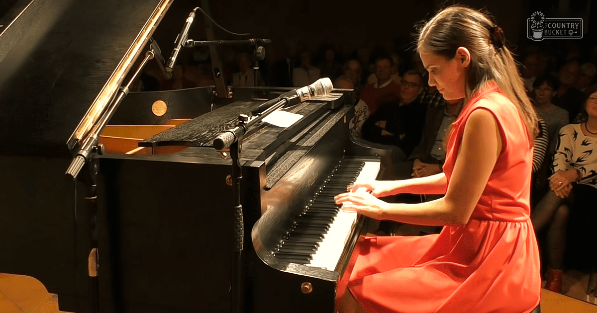 A young adult woman plays piano in a concert hall with an audience seated in the background.