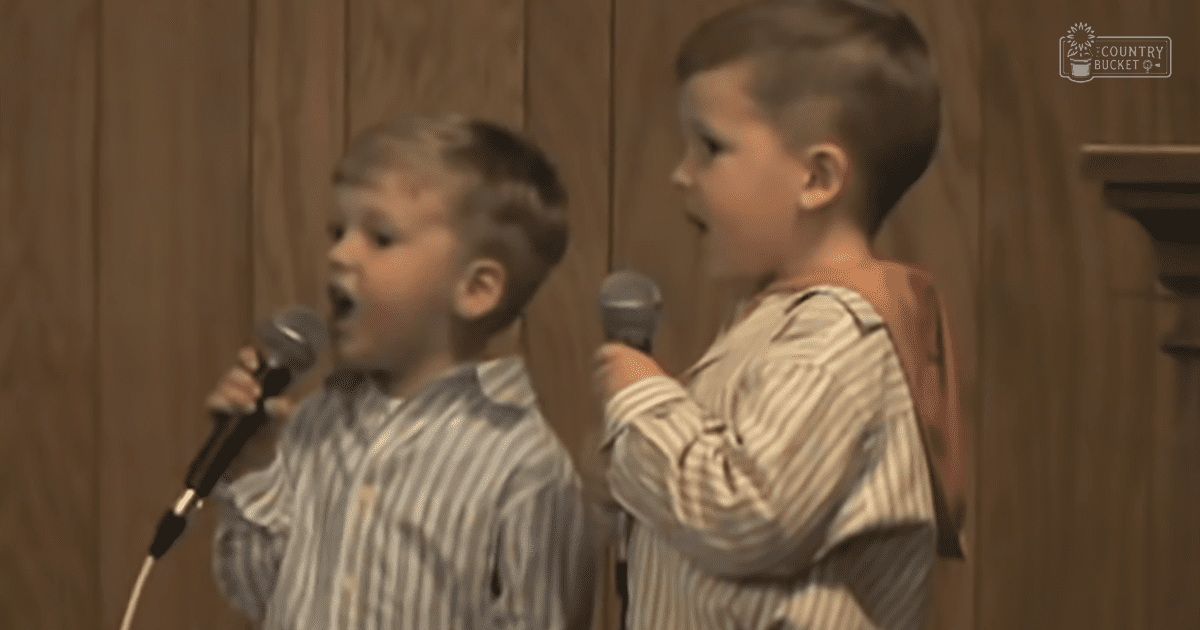 Two young male singers wearing striped shirts perform in a room with wooden paneling.