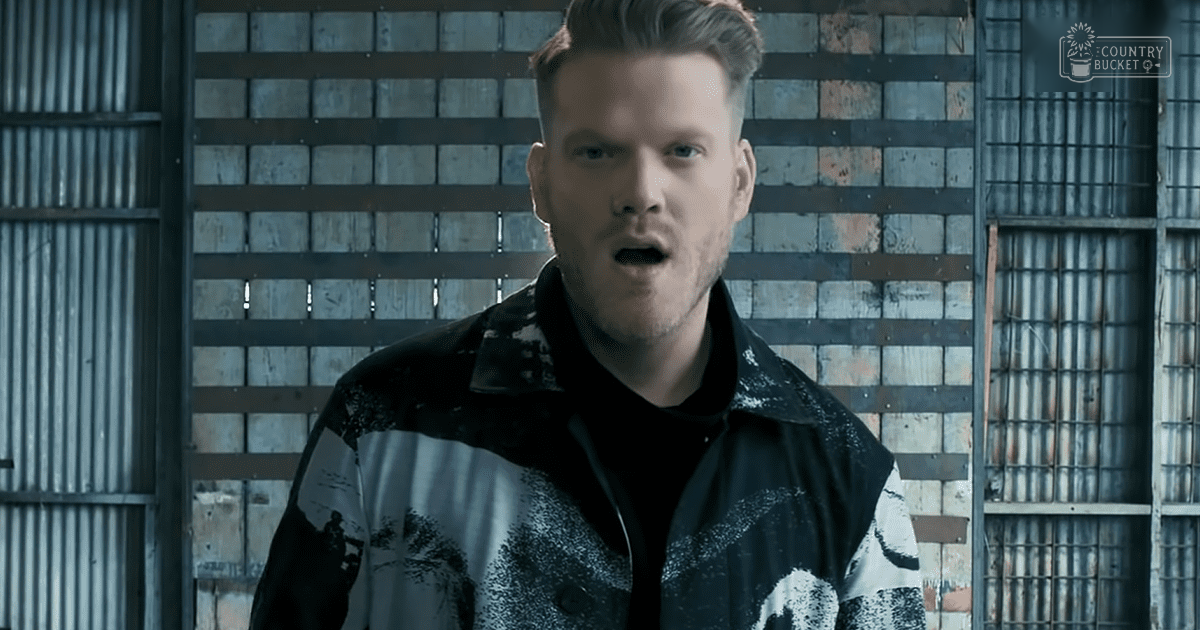 A young adult male in a relaxed fit long-sleeved shirt or jacket with an abstract or graffiti-like pattern stands against metal walls resembling industrial storage units.