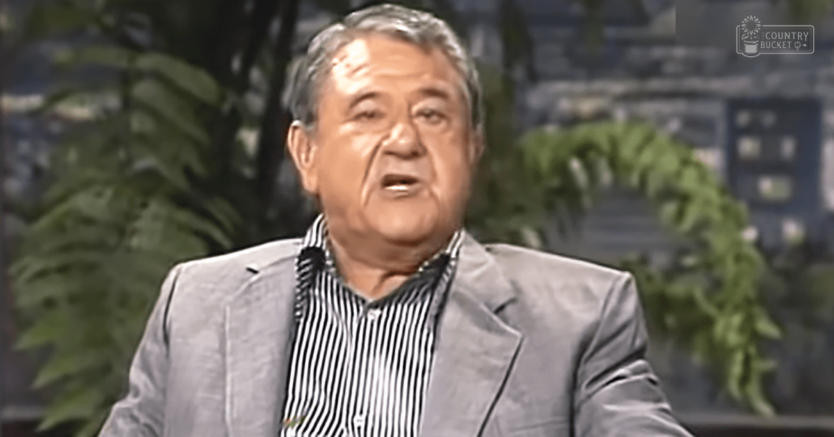 An older adult male wearing a formal gray suit with black and white striped button-down shirt, standing indoors in what appears to be a television studio setting.