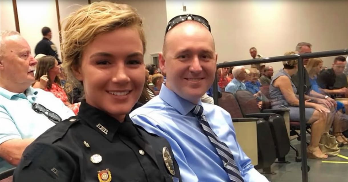 An adult male and a female police officer seated together in a public meeting room or conference hall, with rows of chairs and an audience seated behind them, suggesting a formal gathering or meeting; the scene is well-lit, indicating daytime or early evening.