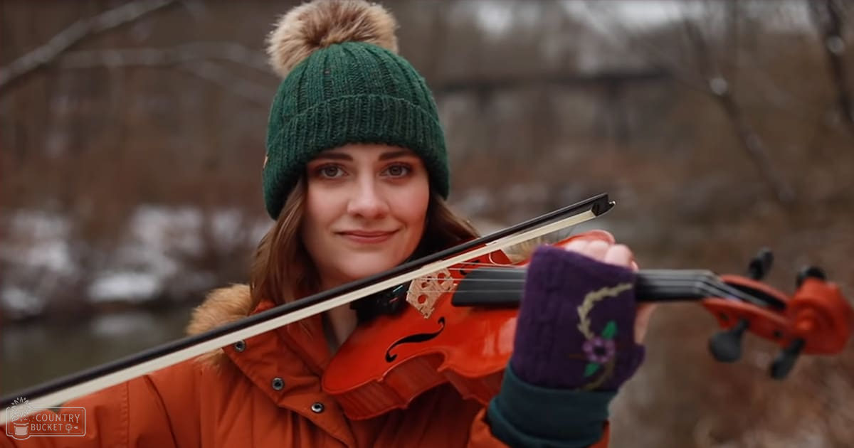 A young adult female wearing an orange jacket with a fur-lined hood and button details, playing a violin outdoors during what appears to be winter or early spring.