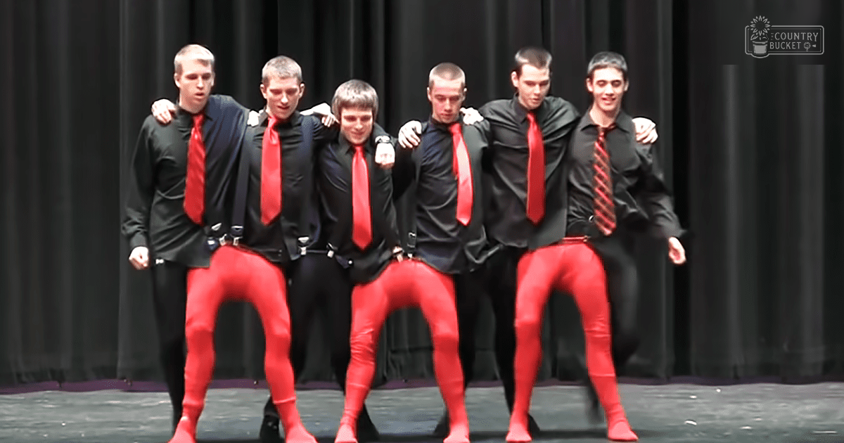 A group of young adult male performers dressed in formal black and red attire stand on stage under bright artificial lighting against dark curtains, suggesting a school play or talent show performance.