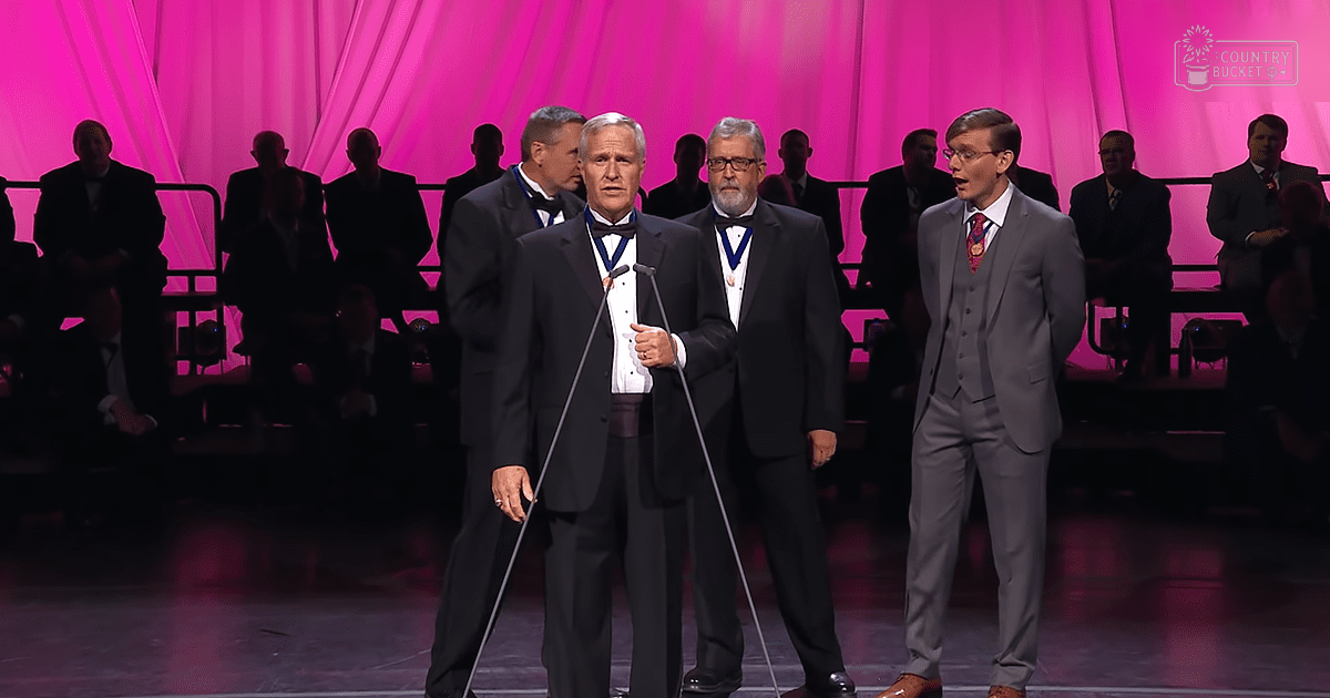 Three older adult males in formal attire present at what appears to be an awards ceremony, standing in front of a stage with a pink curtain backdrop.
