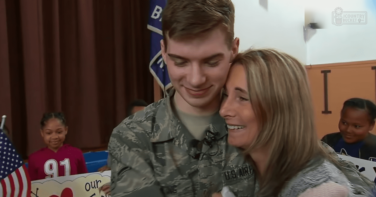 A young male soldier in U.S. Air Force uniform embraces a middle-aged woman with shoulder-length blonde hair, standing indoors within what appears to be a school setting during an event or assembly related to military appreciation.