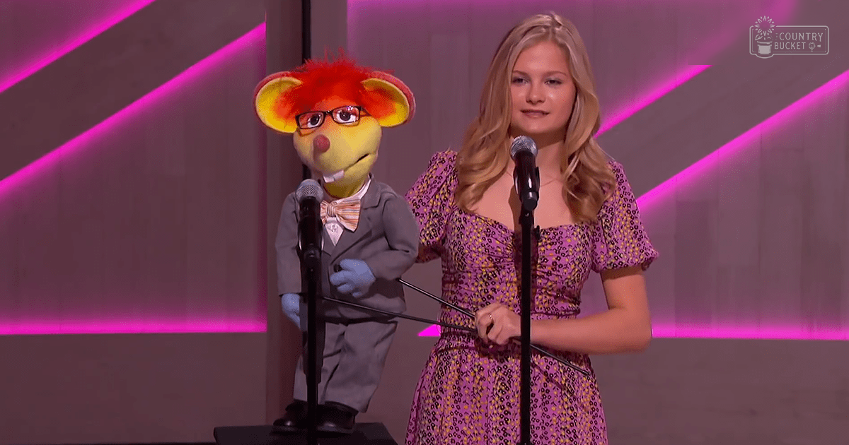 A young adult woman in a purple floral dress performs on stage with an anthropomorphic animal puppet wearing glasses and a gray suit at what appears to be an awards show.