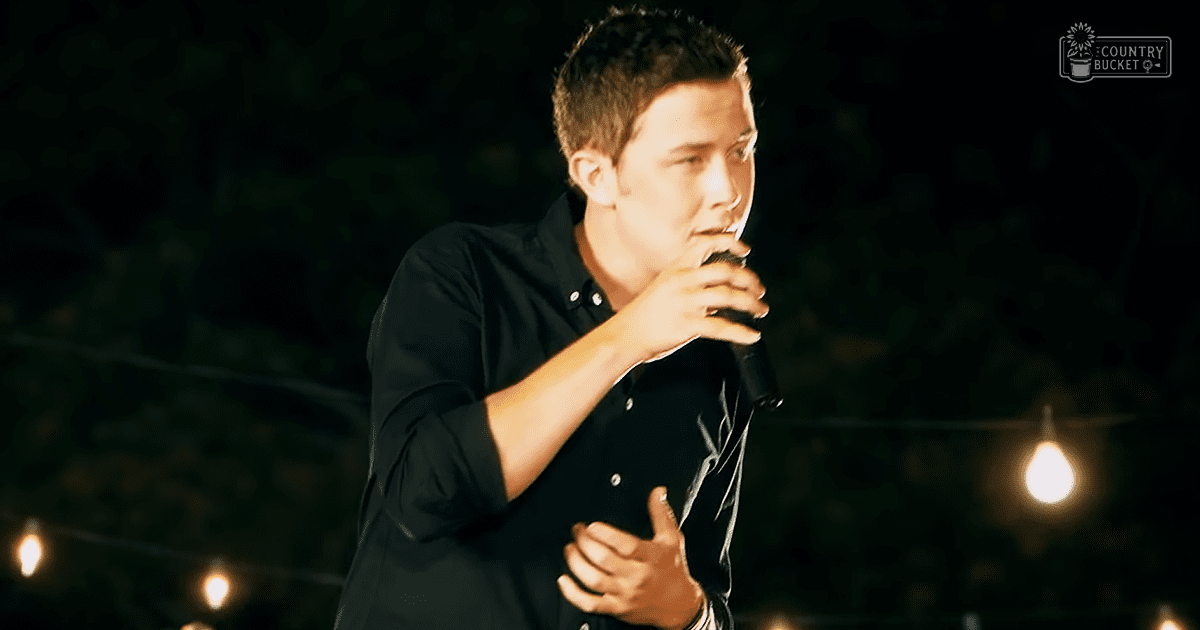 A young adult male singer performing on stage at night under ambient string lighting in a performance venue.