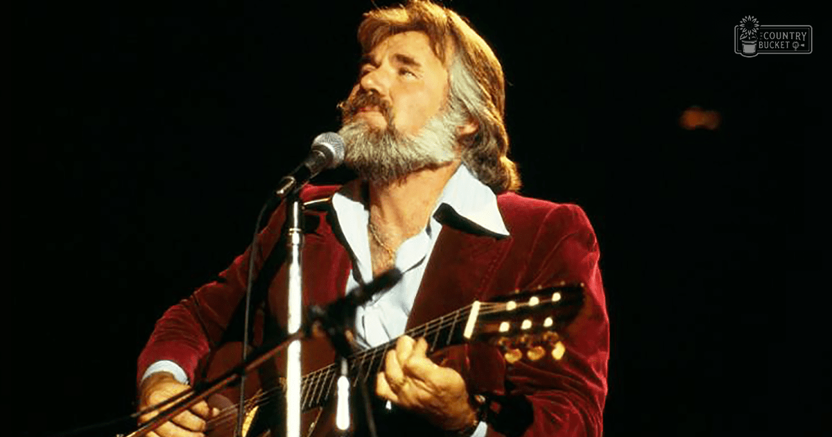 A middle-aged male singer in a red velvet jacket over a white collared shirt with black trim, holding an acoustic guitar on stage under focused lighting.