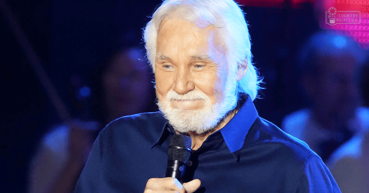 An older adult male in a dark blue, modern-era shirt at what appears to be a concert or event setting with blurred stage lighting.