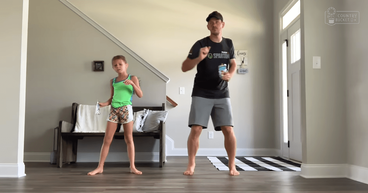 A father and daughter dance indoors, surrounded by home furnishings like a bench with cushions and a checkered rug. The setting appears to be during daytime in their living room or entryway.