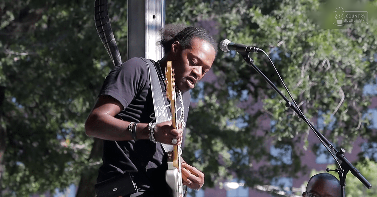 An adult male musician performing outdoors in an urban setting, wearing a dark gray T-shirt with a white design, black pants, several bracelets, and a necklace.