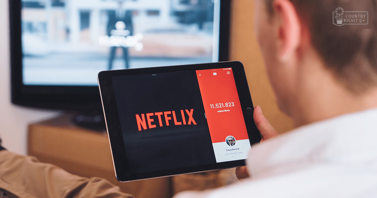 Adult male in a relaxed white shirt using a tablet displaying Netflix indoors, likely in a living room with soft lighting.
