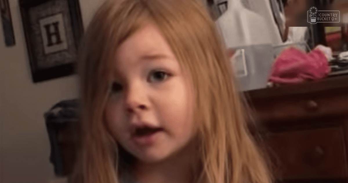 A toddler girl with long blonde hair wearing a light-colored, possibly cotton garment indoors in a home setting. A wooden dresser is partially visible with some items on top.
