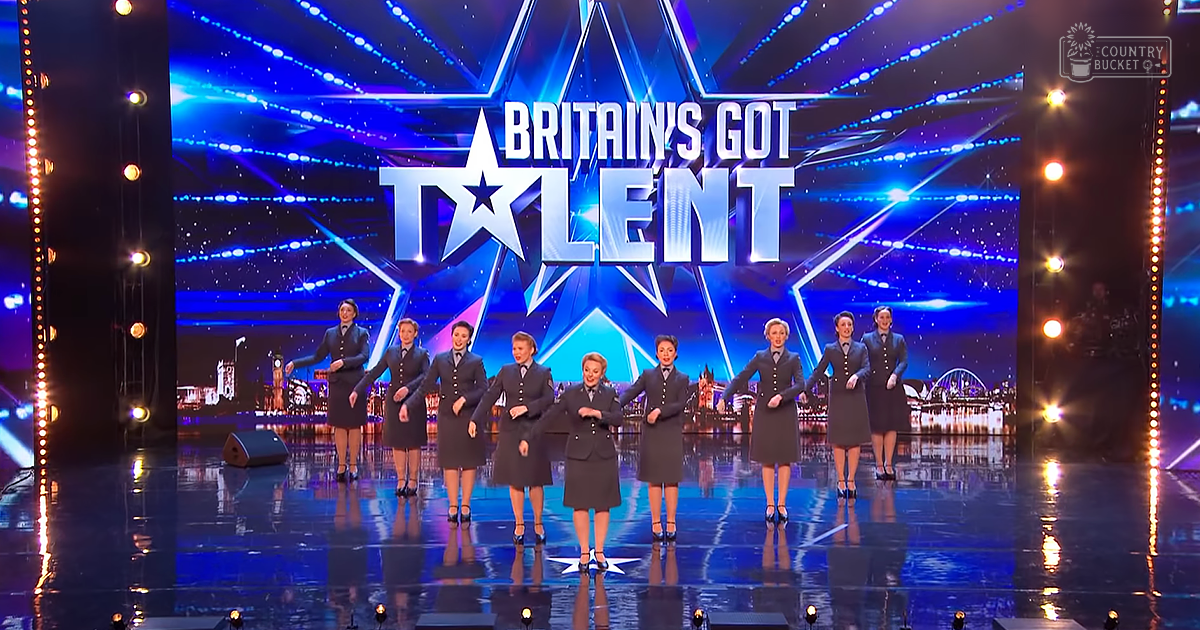 A group performing on stage at 'Britain's Got Talent' in a studio setting, with adults wearing dark gray uniforms inspired by mid-20th century military or airline stewardess styles under bright artificial lighting.