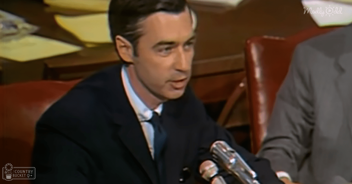 Adult male wearing a dark suit, light blue shirt, and tie at what appears to be a formal indoor event with microphones and wooden paneling.
