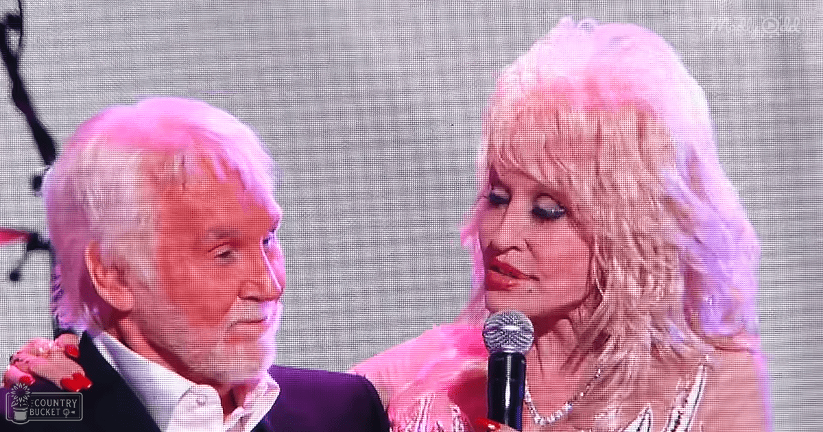 An adult male singer in a dark suit jacket and formal shirt on stage with an adult female performer wearing a shiny metallic pink outfit and pearl necklace, set against a plain backdrop under stage lighting.