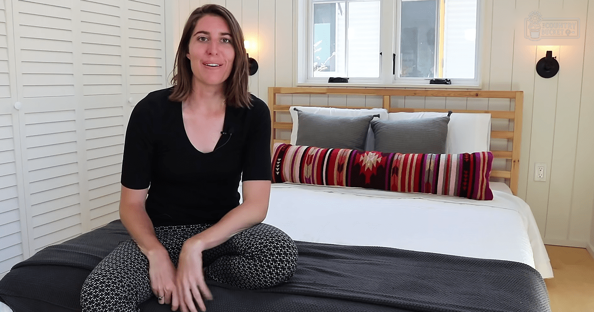 Young adult female in a bedroom setting with a cozy decor style with white walls, brown wooden headboard, gray blanket, colorful patterned pillow, and natural light.