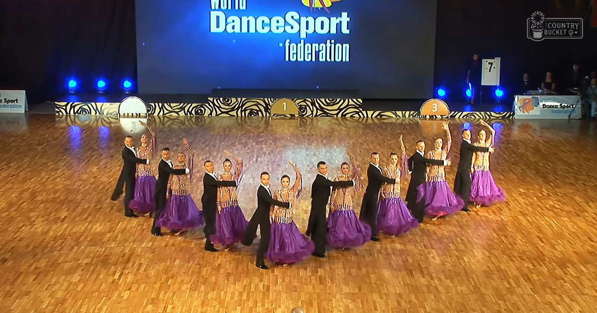 A dance competition scene at an indoor venue with male dancers in black suits and female dancers in purple dresses, set on a polished wooden dance floor under bright artificial lighting.