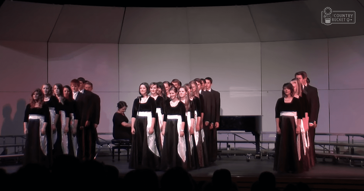 A choir performance in an auditorium with young adults dressed in flowing black and white dresses, seated on benches under artificial stage lighting.
