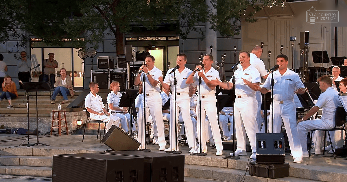 Young adult male performers singing at an outdoor concert venue with trees providing shade and a stage setup.