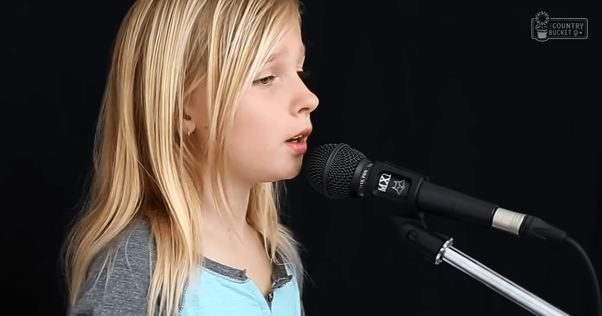 A child singing into a professional microphone, wearing a light blue long-sleeved shirt with darker blue accents and a V-neck.