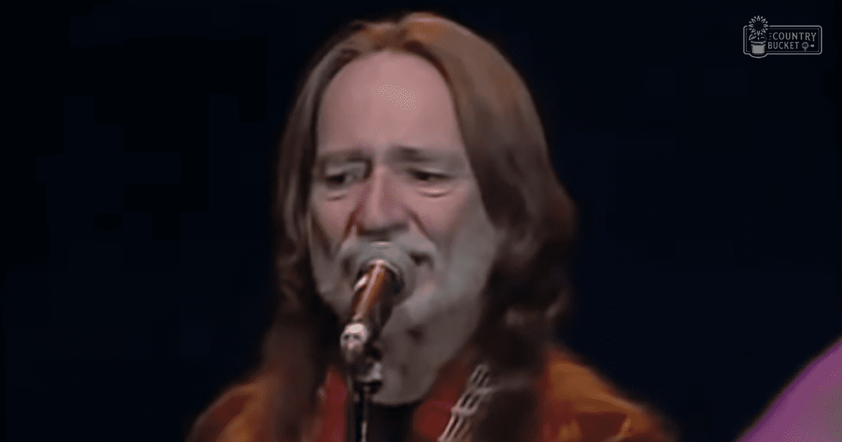 A middle-aged male musician wearing an orange tie-dye shirt performs on stage with a microphone and guitar, under concert lighting at Madly Odd.