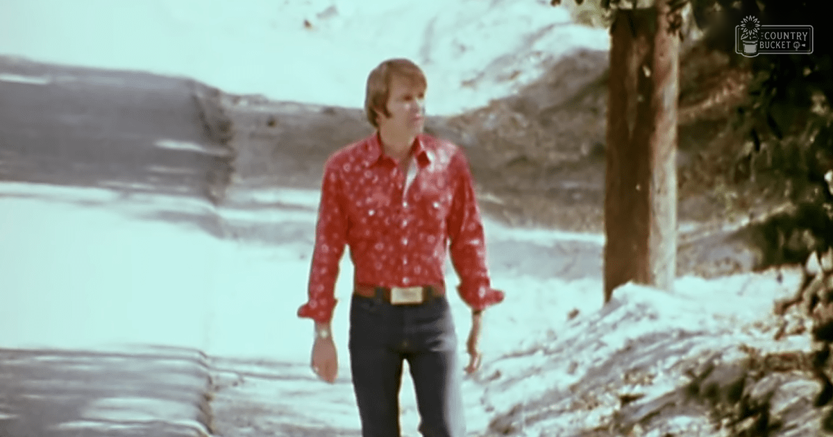 A young adult male in a red patterned shirt with white dots and dark pants stands outdoors amidst snow-covered terrain, wearing a watch on his left wrist.
