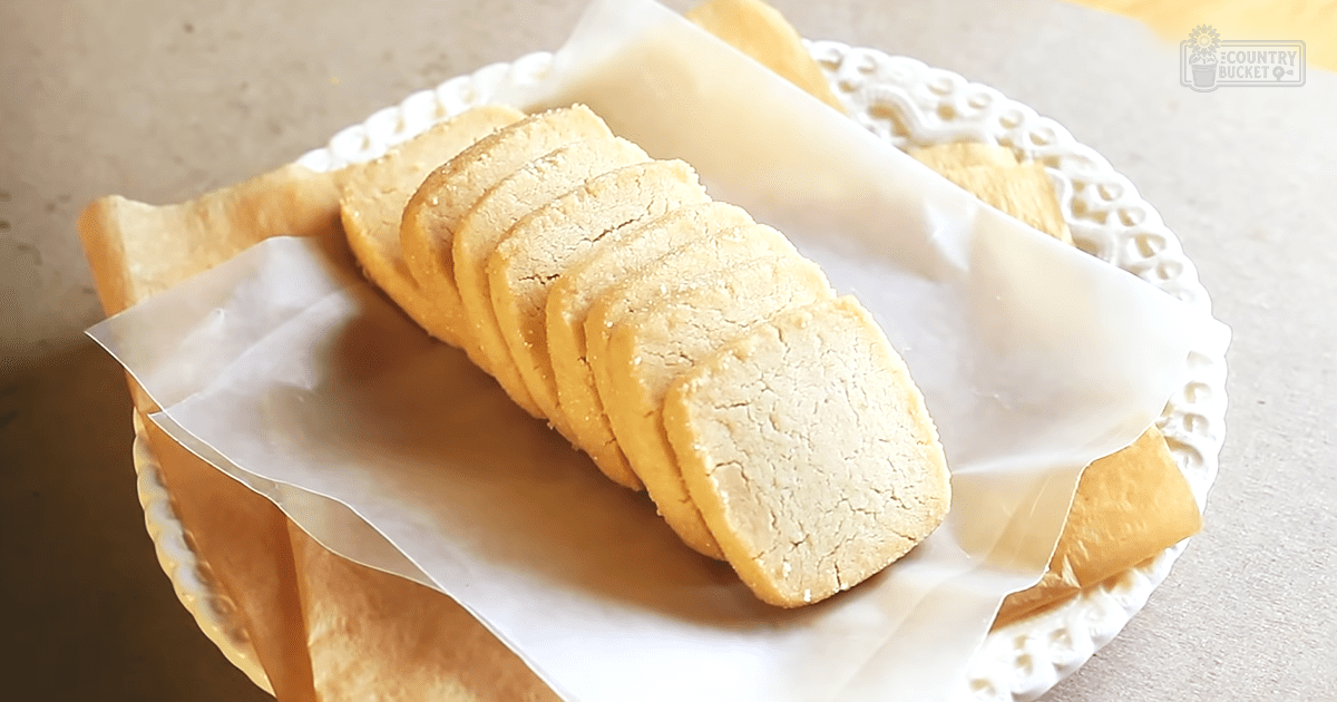 A close-up shot of a white lace-edged plate with parchment paper holding several slices of golden-brown, sugar-dusted bread in an indoor kitchen setting.