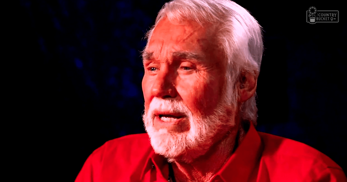 An older adult male wearing a red shirt in an indoor setting with dim lighting. Some indistinct shapes suggest trees or foliage behind him.