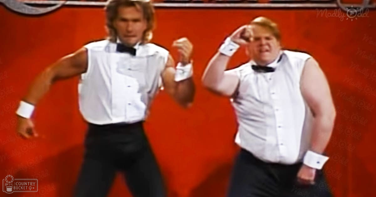 Two adult male dancers wearing white sleeveless vests with black buttons, black bow ties, and white shirts with cufflinks stand against a solid orange backdrop during what appears to be a dance performance scene under bright stage lighting.
