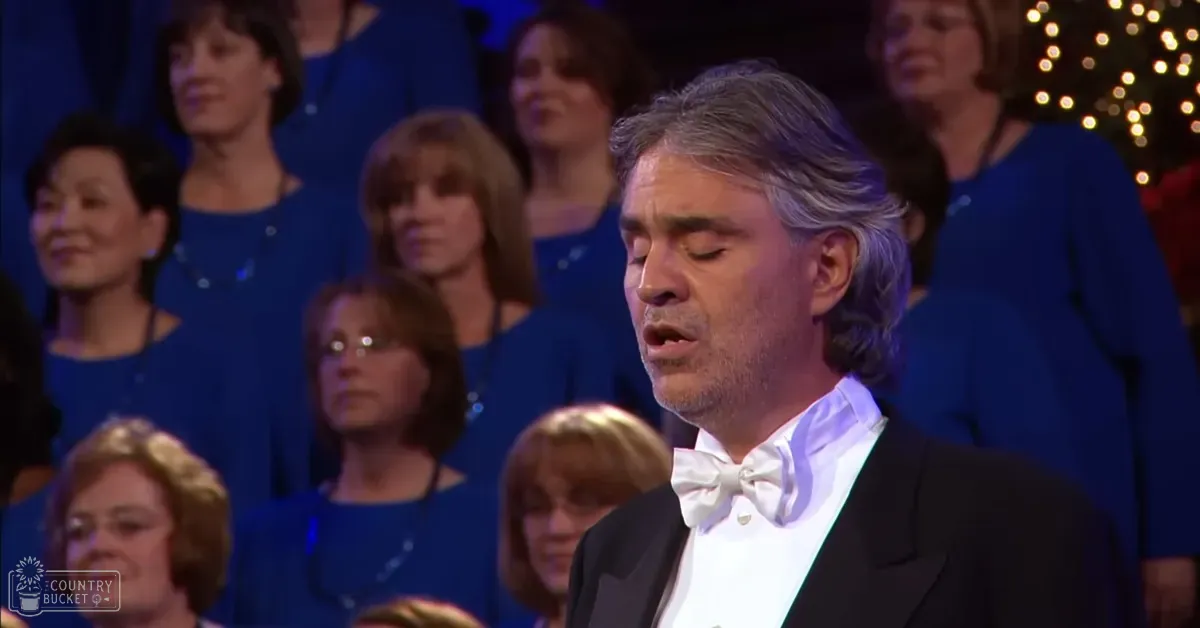 A male adult in a black tuxedo jacket and white bow tie stands on stage, surrounded by a choir of older women wearing blue dresses with necklaces; the setting appears to be an indoor concert hall or theater under stage lighting.