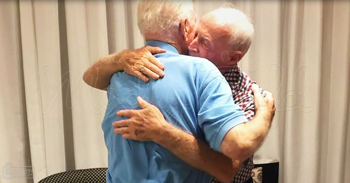 Two older adults embracing each other in a room with vertical blinds, wearing casual attire.