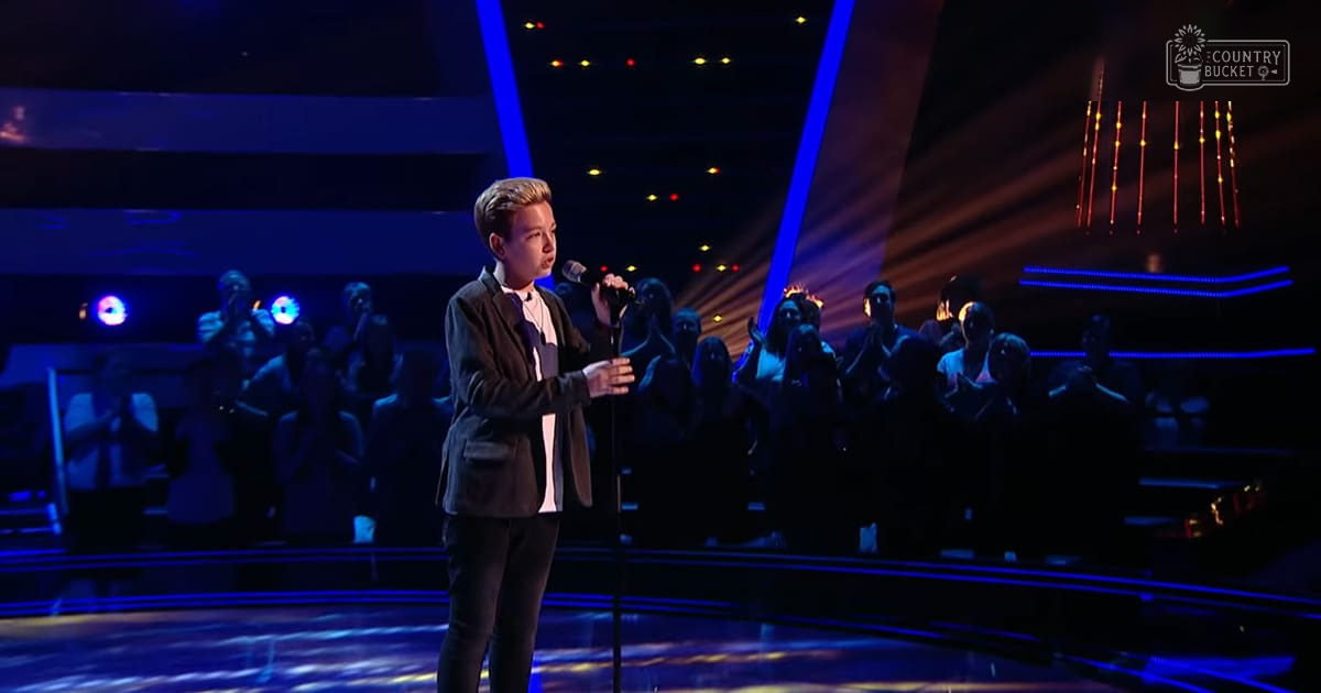 A young adult male singer wearing a dark blazer over a white shirt, holding a microphone on stage with blue and yellow lighting behind him during an evening talent show.