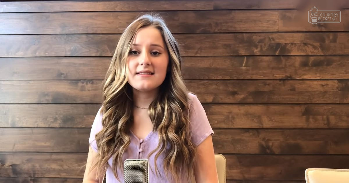 A young adult female wearing a button-down, short-sleeved, light purple or lavender top with soft cotton blend fabric stands in front of a wooden wall setup for an interview or recording.