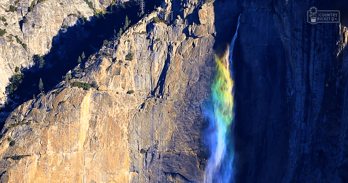 A vibrant waterfall with a rainbow effect in Yosemite National Park under midday or early afternoon light.