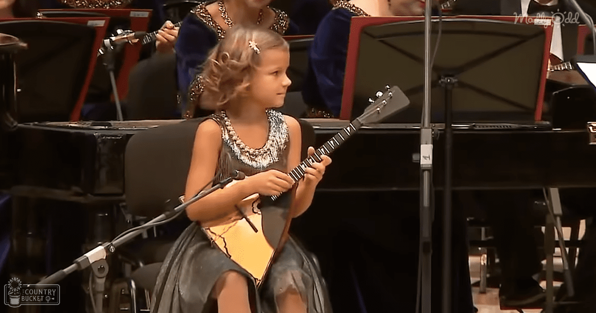 A young girl plays mandolin in a formal indoor orchestra setting, surrounded by musicians and sheet music stands.
