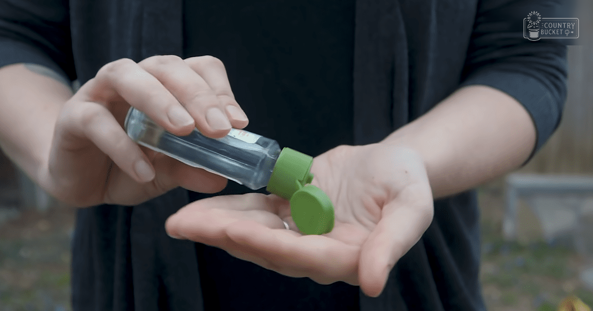An adult female wearing a loose, dark gray long-sleeved shirt uses hand sanitizer outdoors in a garden setting with natural light.