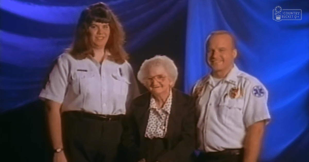 An older adult woman in a formal setting with blue drapes as the backdrop, surrounded by uniformed individuals.