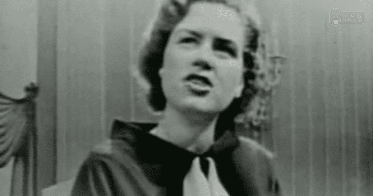 A middle-aged woman wearing a dark, fitted blouse with wide sleeves stands indoors against a wall with vertical paneling or curtains.