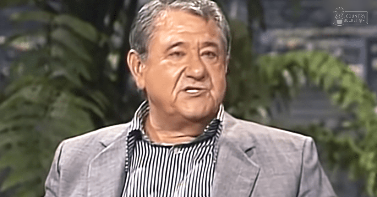 An older adult male wearing a light gray blazer over a black and white striped shirt, set against indoor greenery with 'Madly Odd' watermark.