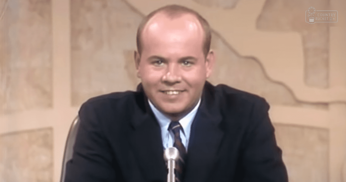 Middle-aged male in a formal dark suit with a light blue dress shirt and patterned tie, seated against a textured stone wall with a microphone nearby, suggesting an indoor setting.