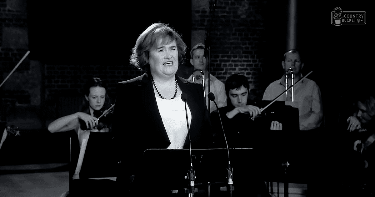 A woman singing in front of an orchestra at a concert with exposed brick walls, stage lighting, and microphones visible.