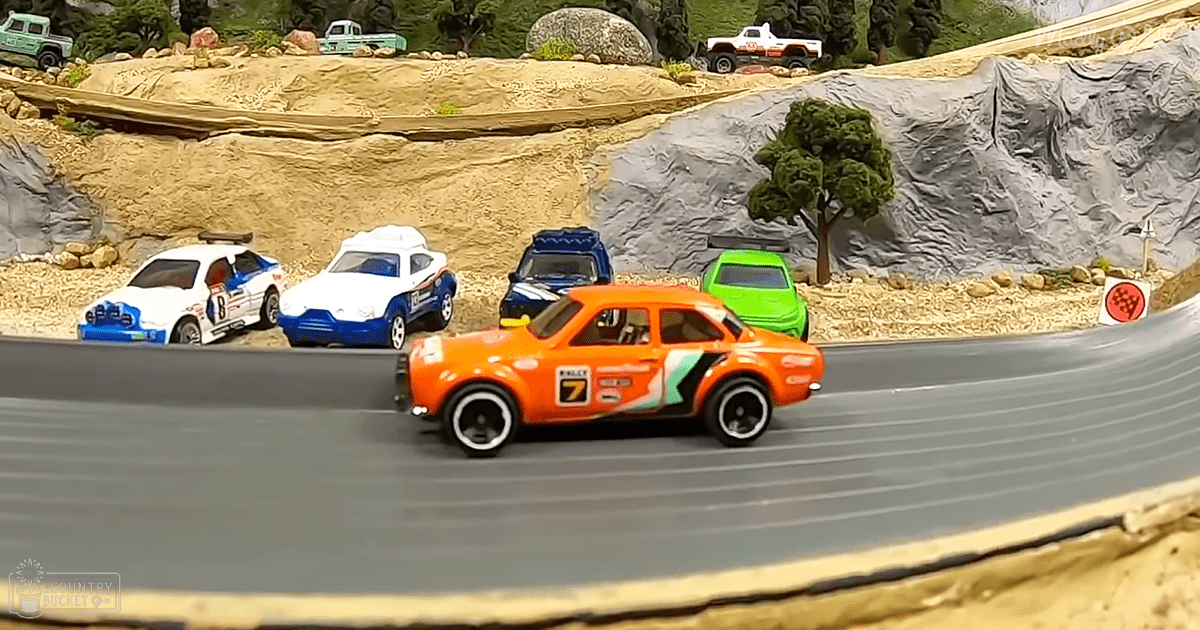 A detailed diorama model of a toy car race track with orange foreground car under bright daylight.