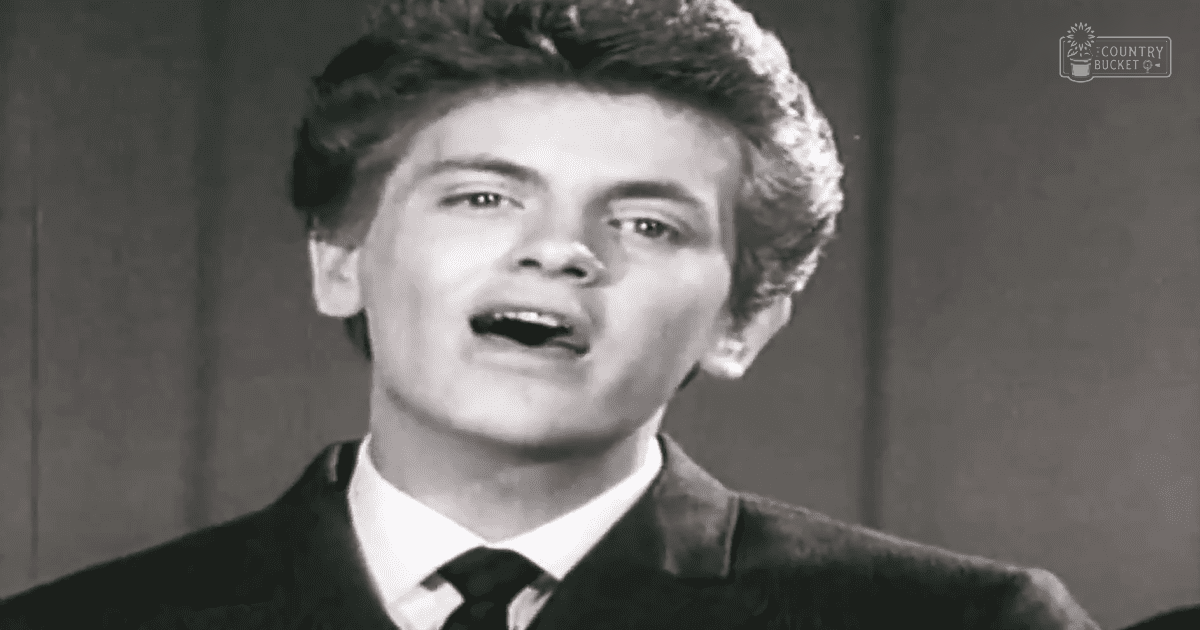 A young adult male in a mid-20th century-style dark suit jacket stands against an indoor paneled wall with simple lighting, possibly daytime or early evening.