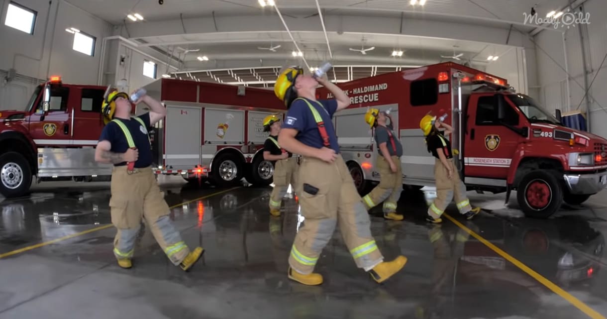 A group of firefighters in a concrete indoor fire station with two red fire trucks parked inside. The scene includes firefighters wearing navy and dark blue t-shirts with reflective stripes and khaki pants, along with yellow helmets and boots.