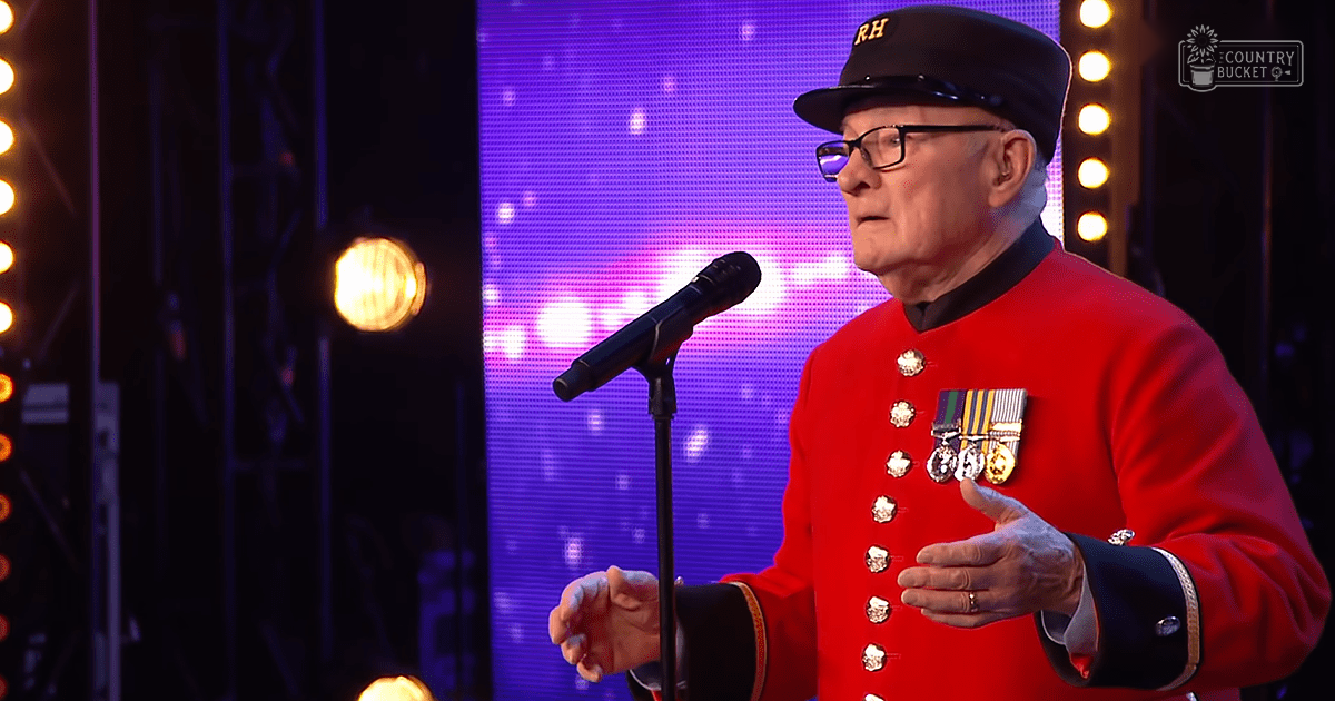 An older adult male in a ceremonial red jacket with medals, wearing a black hat and glasses, performing on stage with a microphone stand and a purple backdrop.