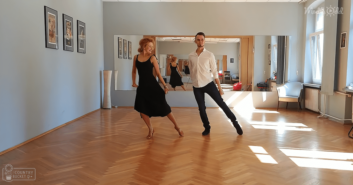 Two dancers in a dance studio scene with wooden floors, natural light streaming through windows, and framed pictures on the wall.