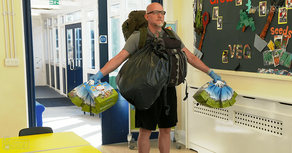 An adult male wearing a casual outfit with gloves, carrying a large backpack and holding plastic bags filled with items in an indoor classroom setting with yellow table and blue doors.