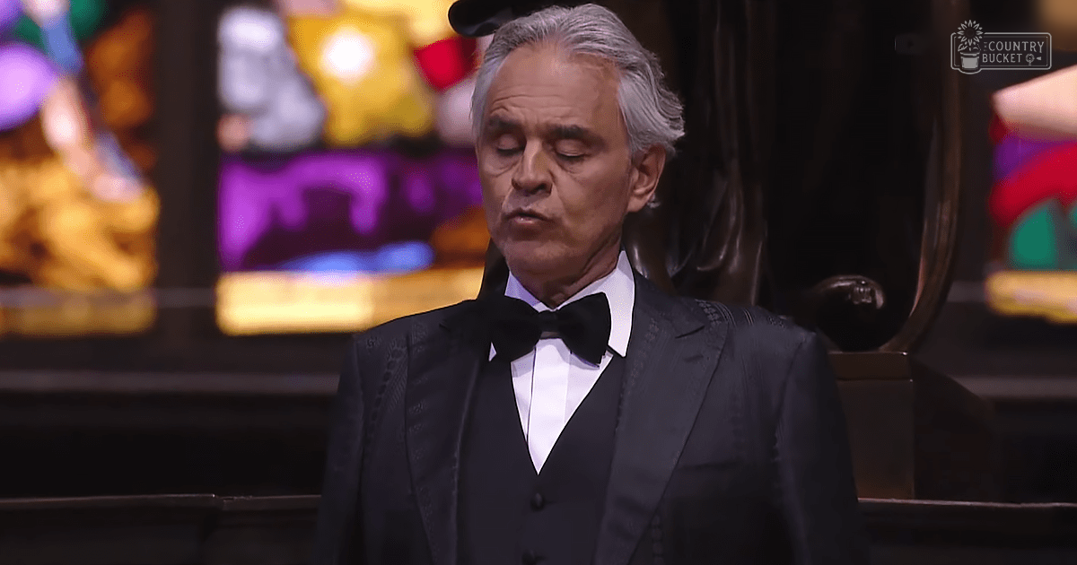 A middle-aged man dressed in a formal black tuxedo with white shirt and bow tie sits at what appears to be an event or performance venue, surrounded by colorful stage lighting.